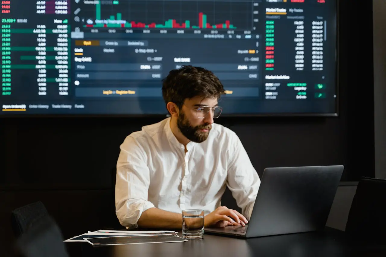 A man in a white shirt sitting at a desk with a laptop, analyzing stock data displayed on a large screen behind him. The screen shows stock charts and figures, symbolizing the complexities and opportunities of investing in foreign stocks, such as Chinese equities, through American Depositary Receipts (ADRs) and US brokerages.